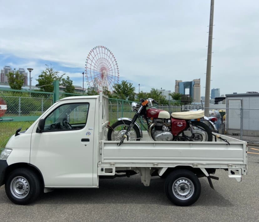 An exterior shot of a vintage motorcycle being loaded onto the back of a small flatbed truck. There's a large ferris wheel in the background so it might be in Yokohama.