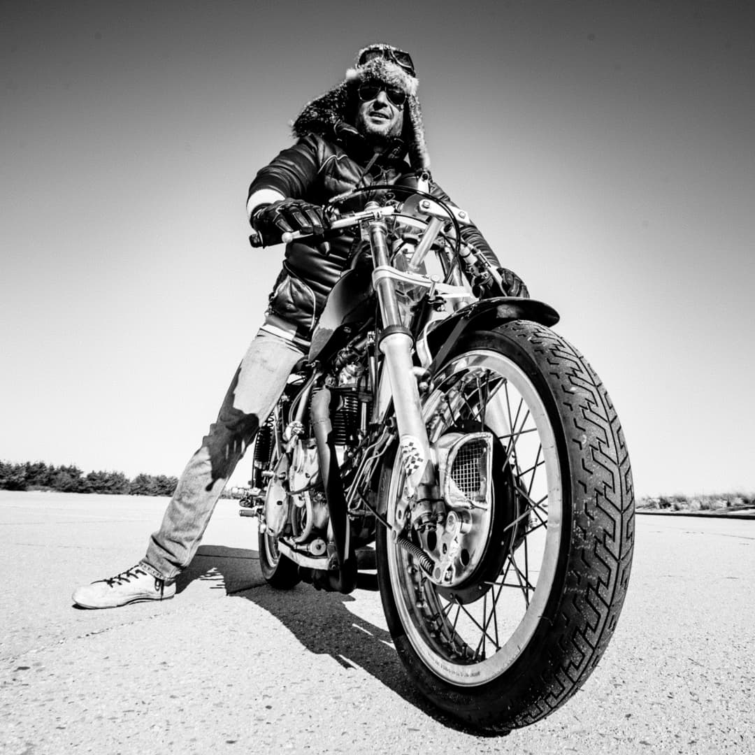 A black and white image of Larry from New York City Motorcycles posing on a vintage motorcycle wearing a fur hat. He is wearing sunglasses and rider's goggles; it's a wonder he can see anything at all.