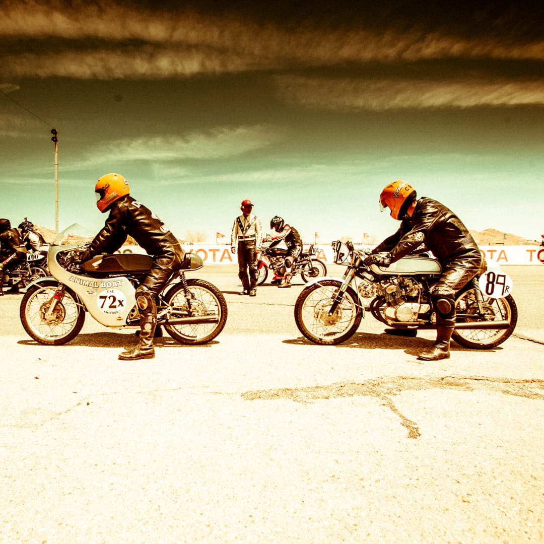 An image of two riders on vintage motorcycles lining up for the start of a race wearing full leathers and orange helmets under a dramatic sky.
