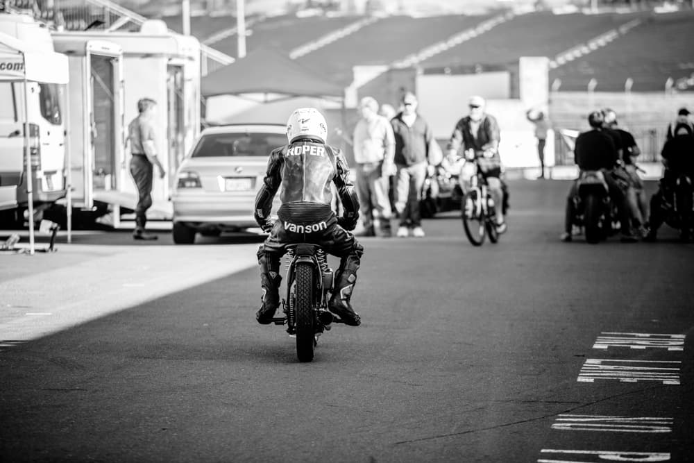 A black and white image a rider in full leathers on a vintage motorcycle. The leathers have the words 'Roper' and 'Vanson' on the back.