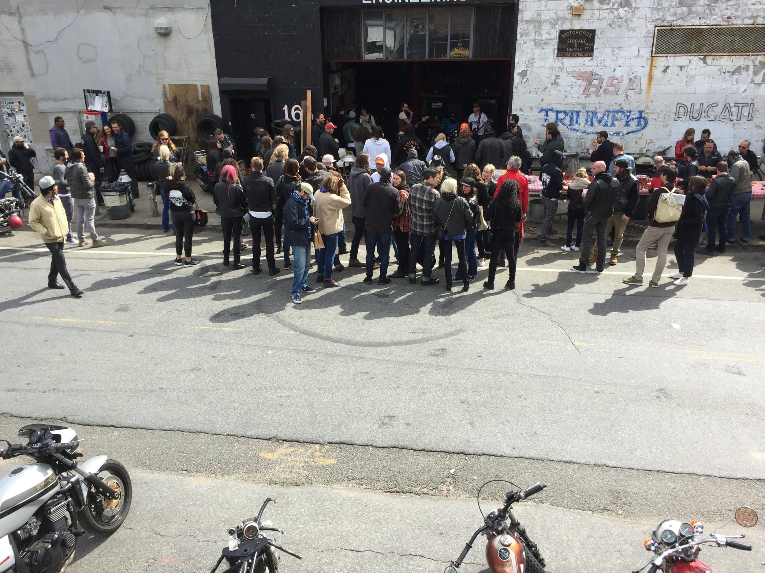 A crowd of vintage motorbike enthusiasts gathered outside the New York City Motorcycles showroom to watch a band that is playing inside.